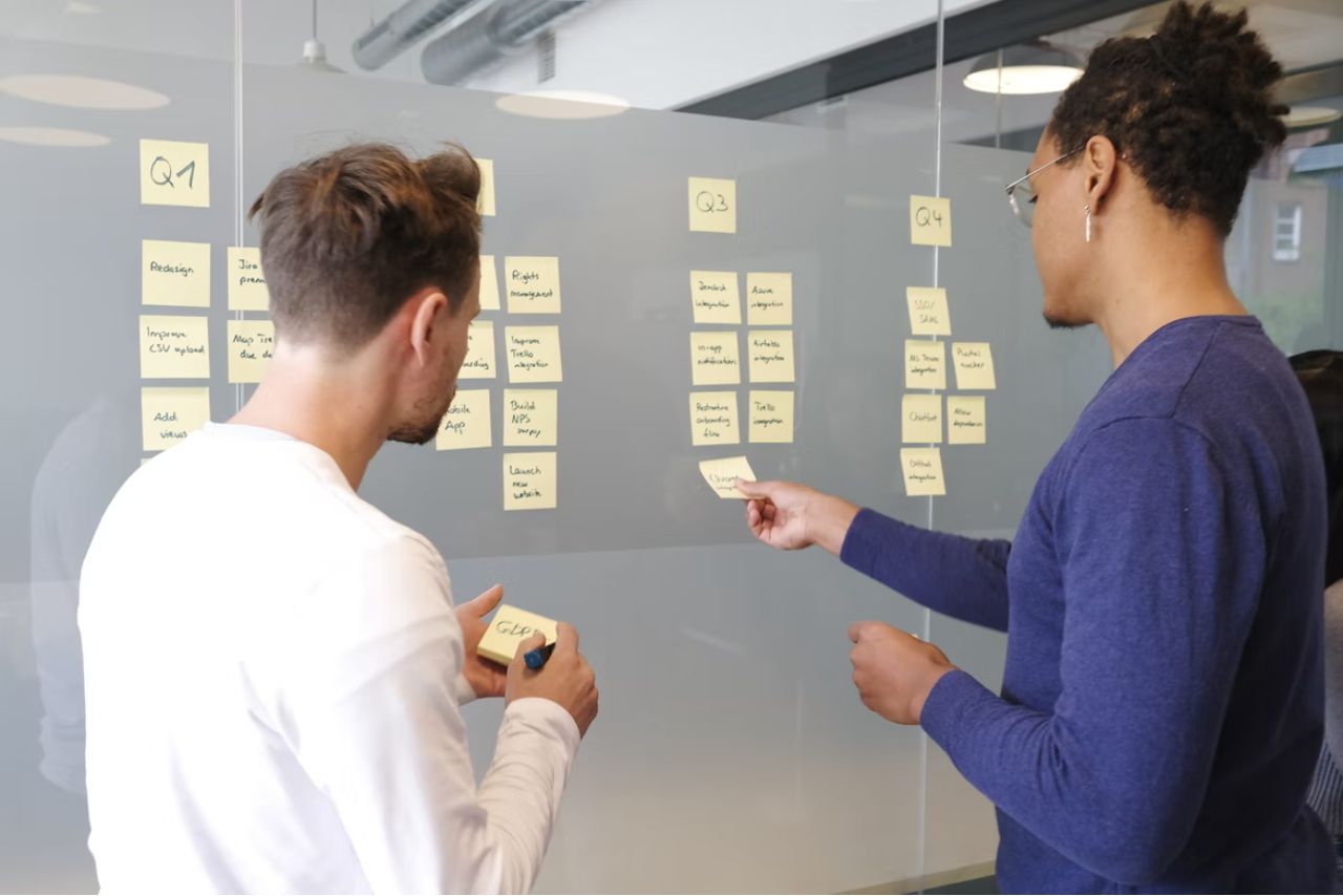 Two young male professionals, one white and one Black, placing post-it-notes on a wall to outline solution architecture goals for Q1 through Q4 of the year.