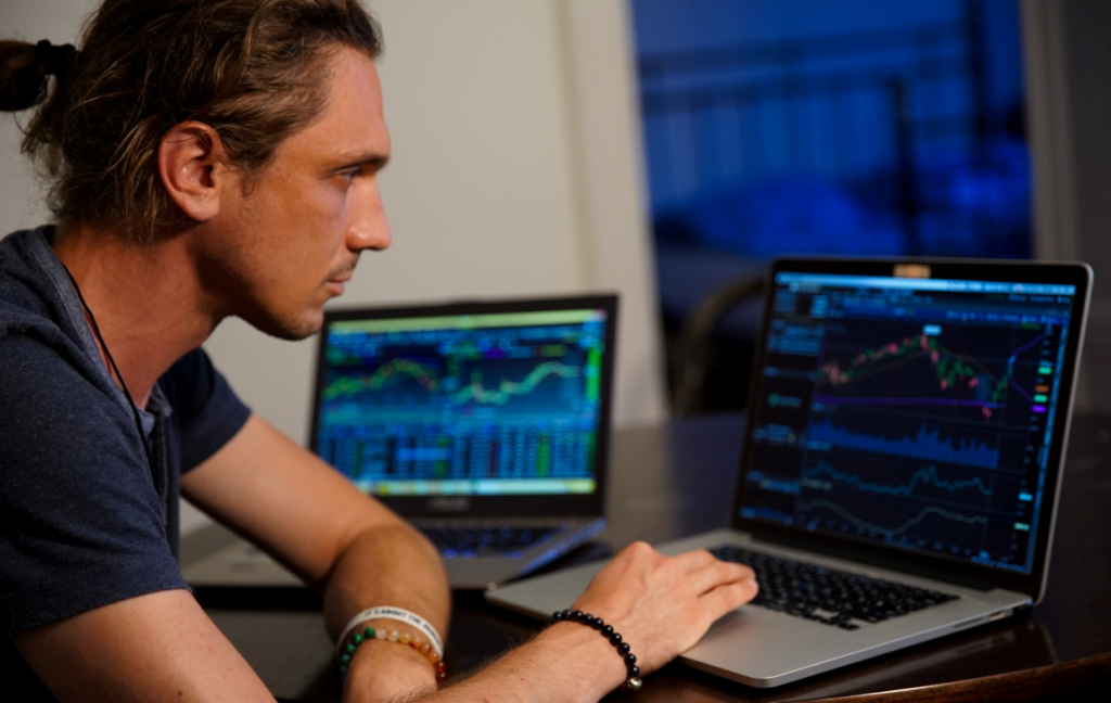 White, male employee in blue shirt with numerous braclets checking data quality metrics on two different laptops