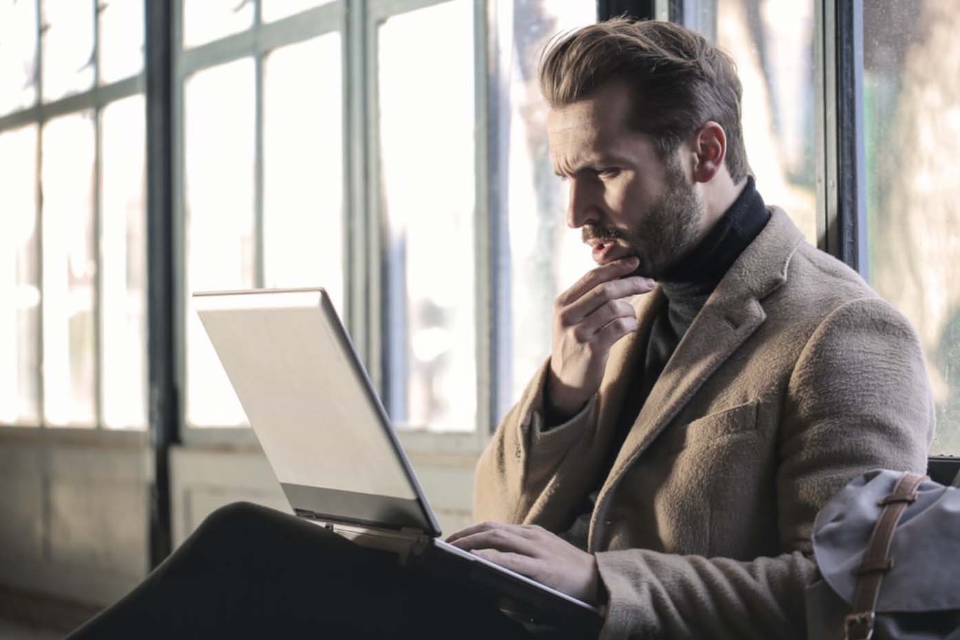 White male professional with slicked-back hair, black turtleneck, and brown coat holding laptop and confused by disorganized code