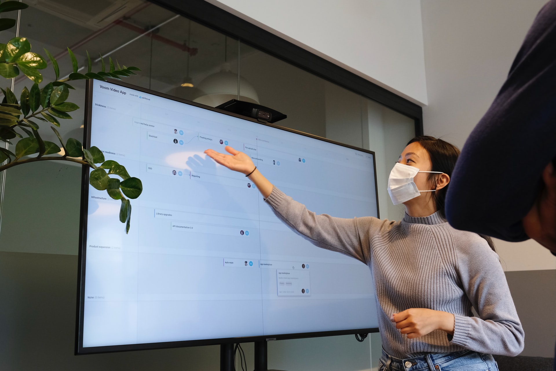 Young Asian woman with grey sweater and white mask holding out her hand to direct viewer's attention to application modernization timeline on television screen