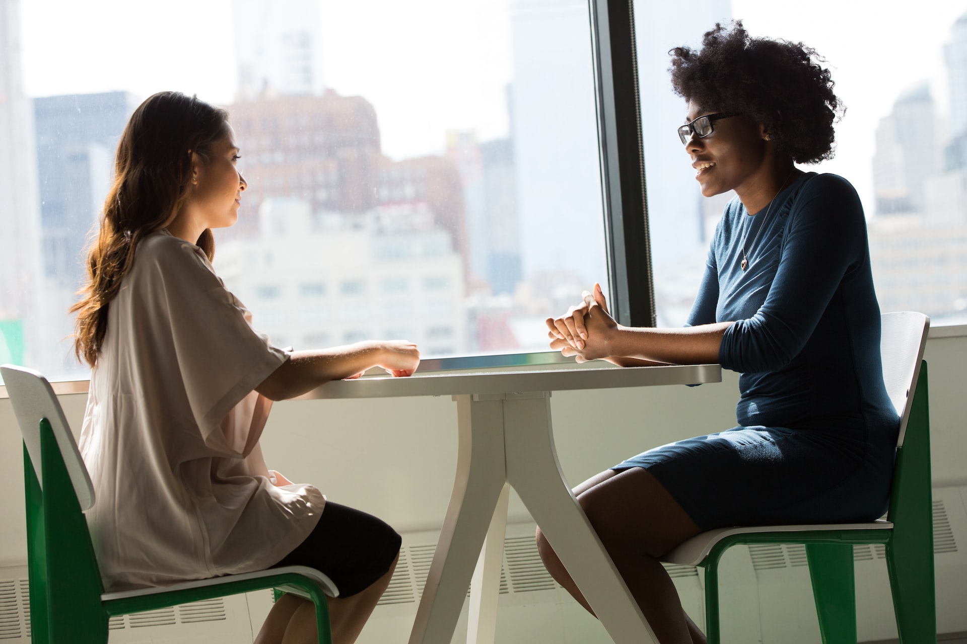Consultation between young Black female in blue sweater and young woman in short-sleeved white shirt together at silver table with a skyline view in the background.