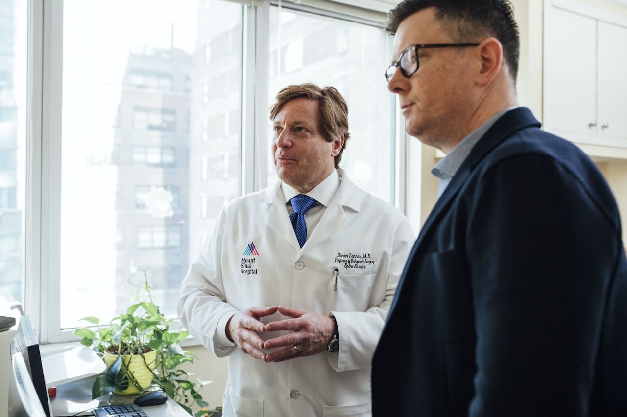 Illustration related to application modernization case study: Middle-aged white male doctor reviewing test results with white male patient with buildings in the background.
