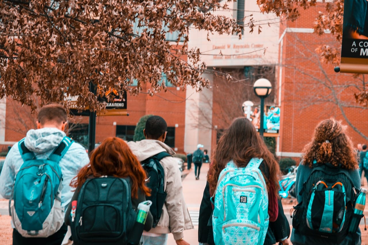 Illustration of application modernization case study: Five young students, three female and two male, with black and blue backpacks walking towards their college campus.
