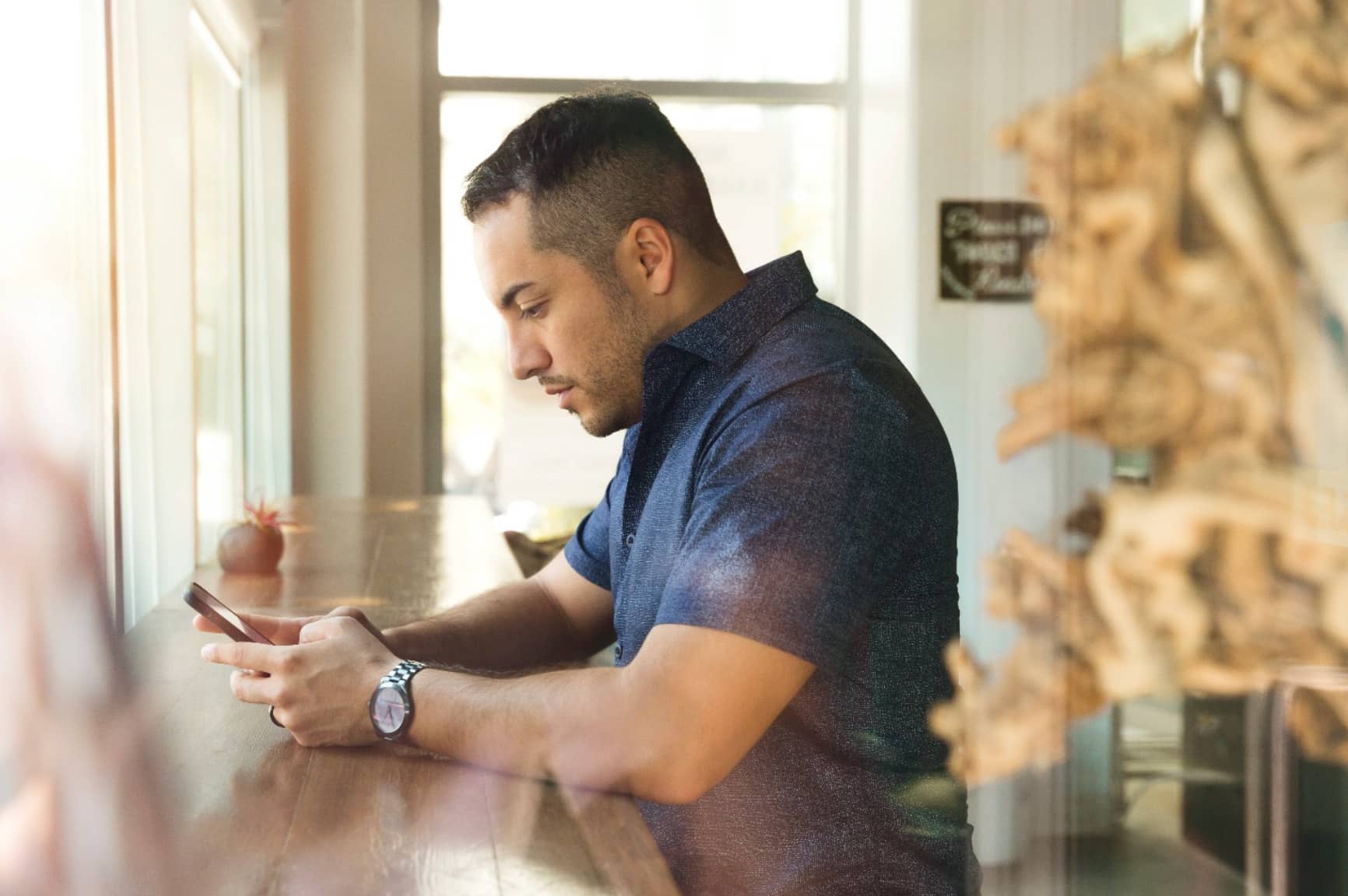 Man in blue button-down shirt with watch on checking phone at coffee shop counter for alert from worksite
